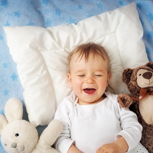 bébé qui rigole juste avant de dormir à côté de deux peluches sur un oreiller blanc