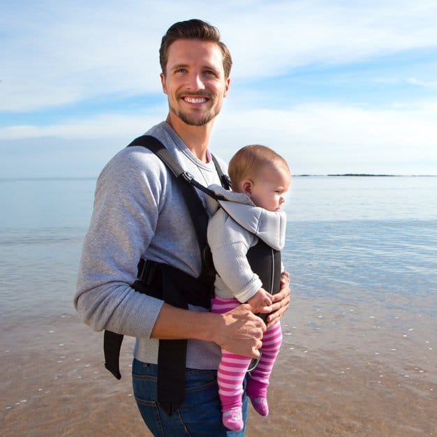 papa qui porte son bébé avec un porte bébé ergonomique à la plage