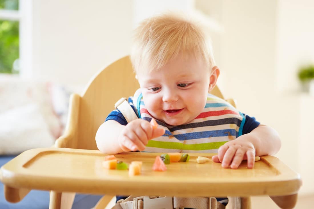 ein Baby sitzt in seinem Hochstuhl und isst in kleine Stücke geschnittenes Obst