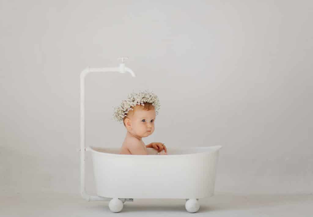 foto von einem babyshooting. ein baby mit einer weißen blumenkrone auf dem kopf sitzt in einer ganz weißen badewanne mit rädern. über seinem kopf befindet sich ein weißer hahn