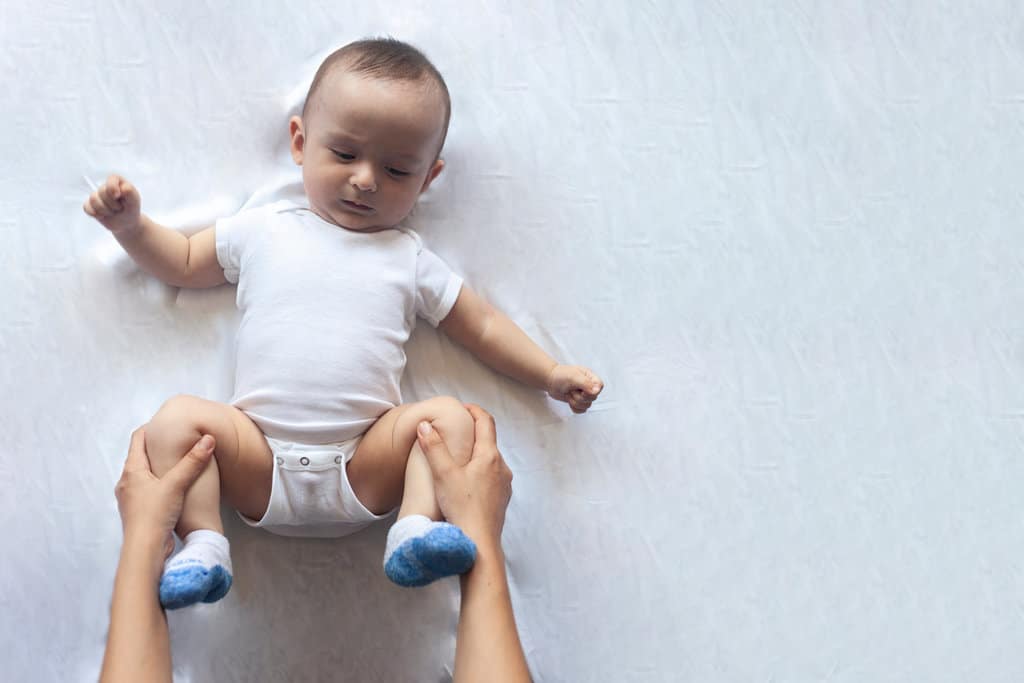 ein Baby liegt mit angewinkelten Beinen und wird von 2 Händen massiert, damit es besser kacken kann