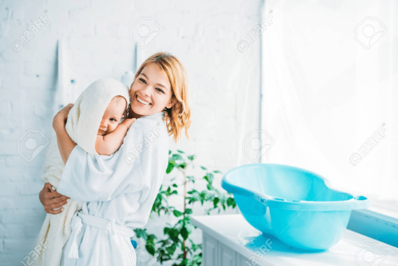 eine Mutter in einem weißen Bademantel trägt ein Baby in ihrem beigen Handtuch, das gerade aus ihrer blauen Badewanne gestiegen ist