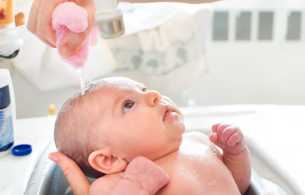 un bébé prend le bain dans une baignoire pour bébé. une main féminine tient sa tête et sa nuque tandis que l'autre main essore un chiffon rose qui fait gouter de l'eau sur la tête du bébé
