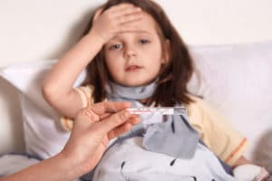 une enfant malade porte sa main sur son front. une main tient un thermomètre avec de la température juste devant la jeune fille.