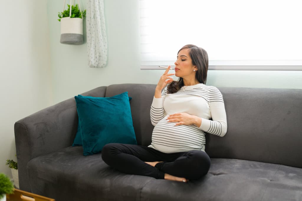 eine schwangere Frau raucht eine Zigarette, sitzt im Schneidersitz auf einem grauen Sofa und geht das Risiko ein, ein hustendes Baby zu bekommen