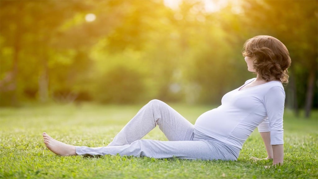 eine im fünften Monat schwangere Frau, weiß gekleidet, liegt im Gras eines von Bäumen gesäumten Parks