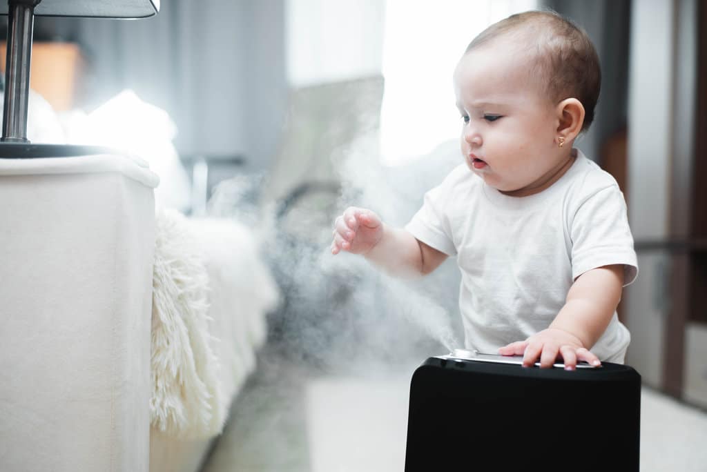 ein Baby in einem weißen kurzärmeligen Body spielt mit einem Dampfbefeuchter in einem Zimmer neben einem Bett