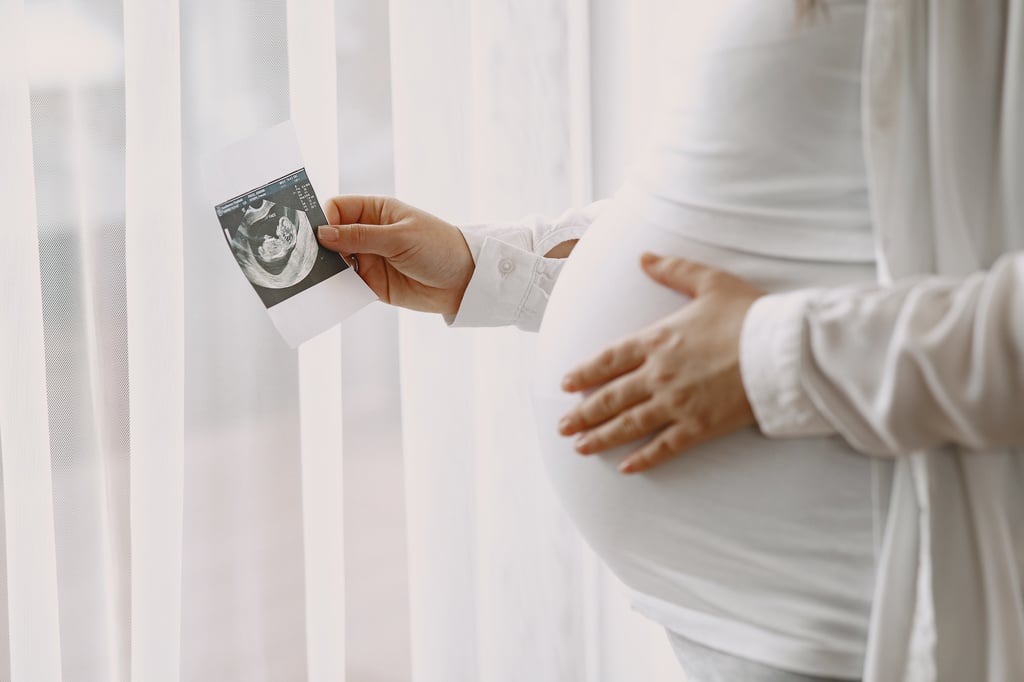 une femme enceinte vêtue de blanc porte une échographie de son bébé dans une main et touche son ventre avec l'autre main